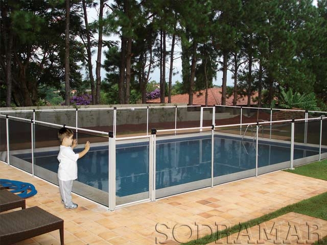 Uma criança vestida de branco fica ao lado de uma piscina cercada em um quintal com pinheiros altos e grama sob um céu nublado.