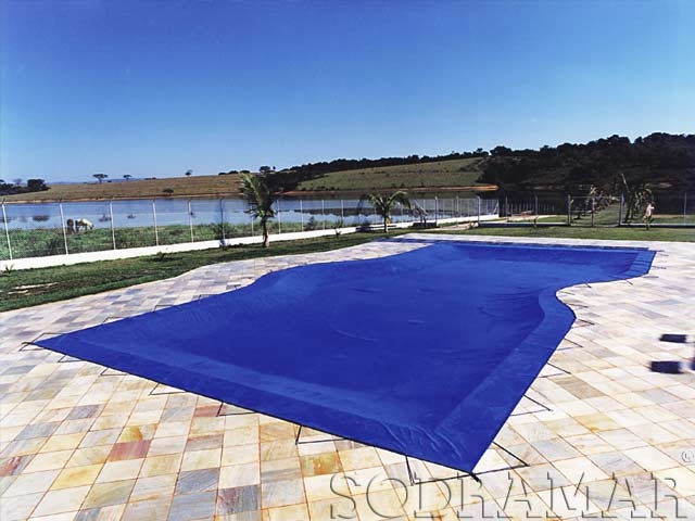 Uma cobertura de piscina sodramar azul estende-se sobre uma piscina de formato irregular, com uma paisagem verdejante e um lago ao fundo sob um céu limpo.