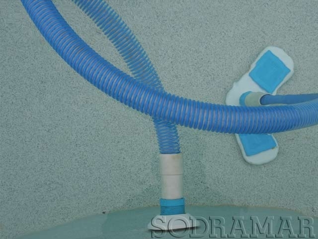 Uma mangueira de limpeza de piscina e um cabeçote de aspirador fixados na lateral de uma piscina azul clara, com a parede cinza texturizada ao fundo. O logótipo "Sodramar" é ligeiramente visível
